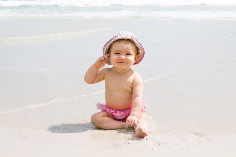 Beautiful little girl is sitting at beach near to sea and playing with sand.