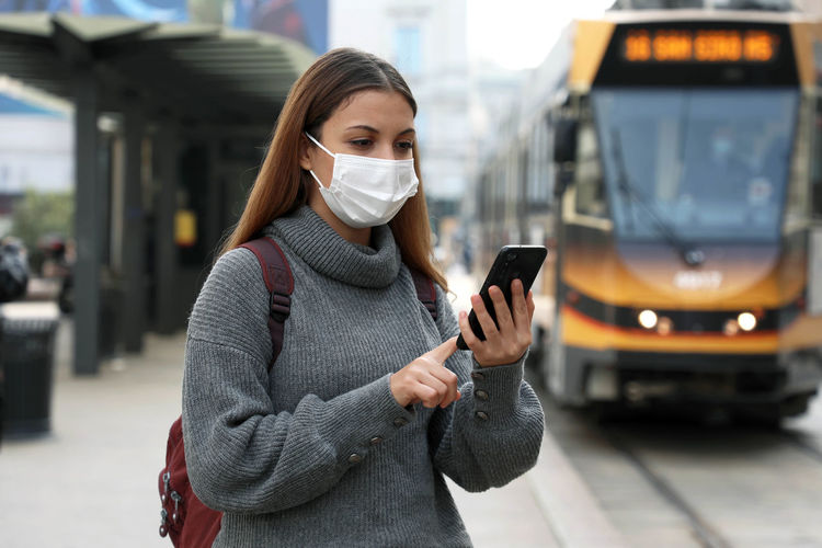 Young woman wearing mask using smart phone while standing on road