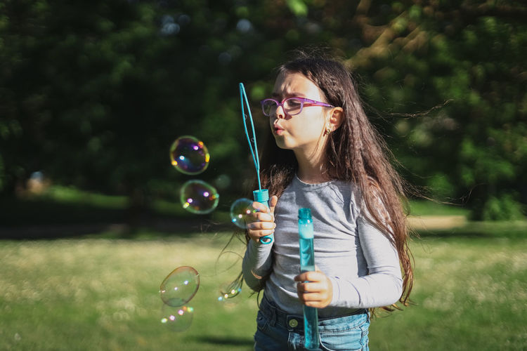 Portrait of a caucasian girl blows soap bubbles while standing in a park on a playground meadow