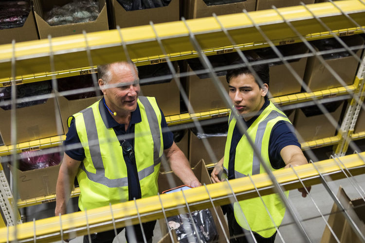 High angle view of male coworkers discussing while seen through rack shelf at warehouse