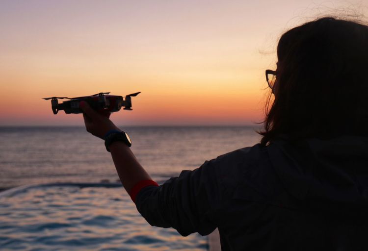 Rear view of woman holding drone at beach against sky during sunset