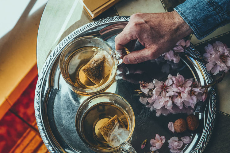 From above hand of crop anonymous man taking glass cup of tea placed on metal tray with flowers in light room