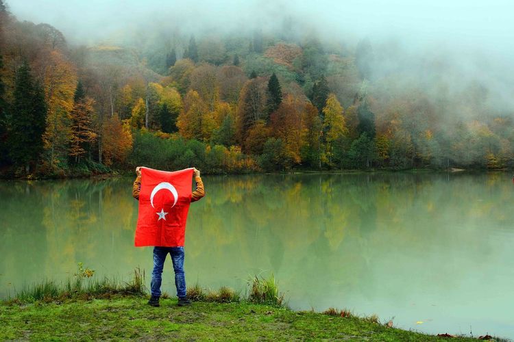 Rear view of man holding turkish flag at lakeshore during autumn