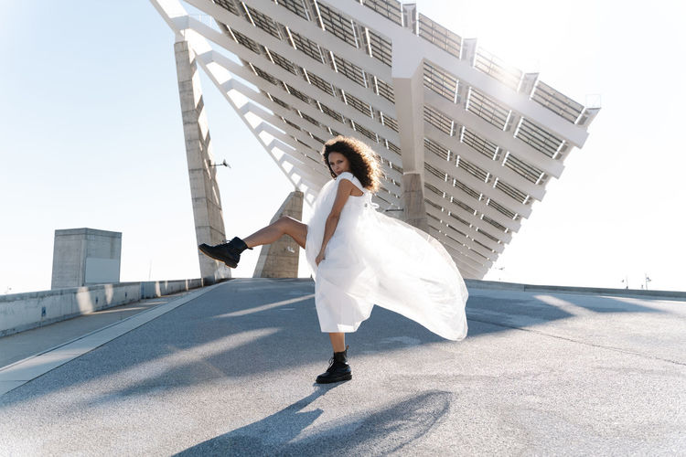 Full body of cool bride in brutal boots dancing looking at camera on paved waterfront with solar batteries on background in back