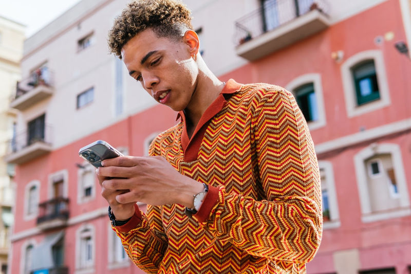 From below side view of young hispanic guy with afro hair in stylish colorful outfit browsing mobile phone while standing on railing near urban building in sunlight