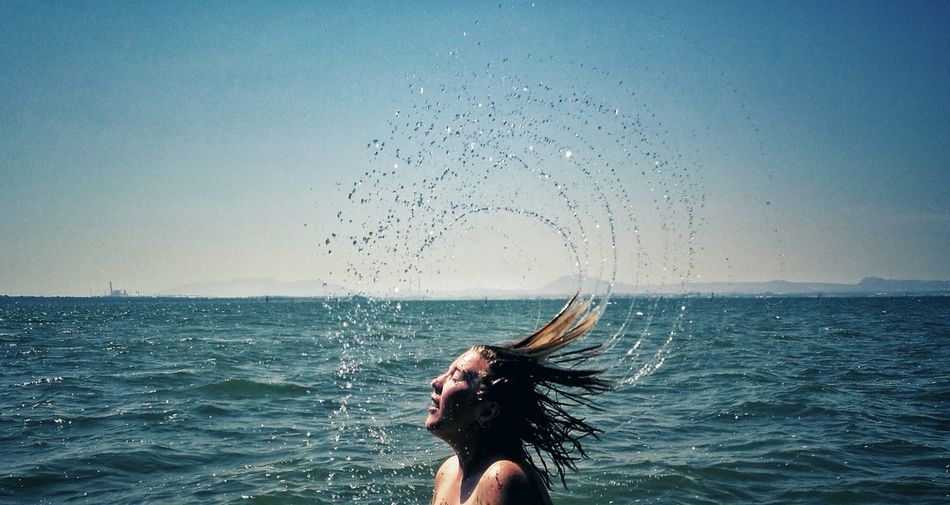 Young woman splashing wet hair in sea against sky