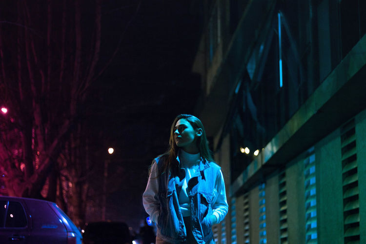 Low angle view of young woman with hands in pockets standing on city street at night