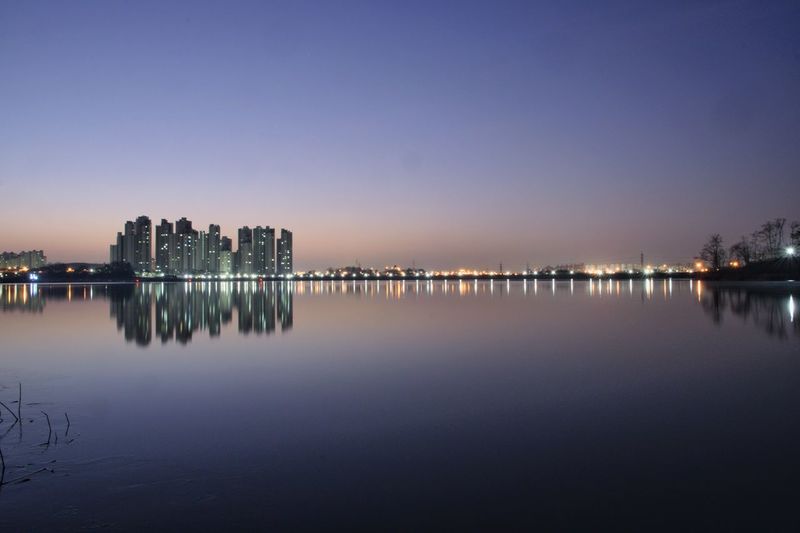 Illuminated buildings by lake against sky at dawn