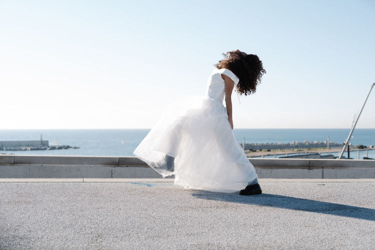 Side view of unrecognizable rebellious female in elegant white dress and brutal boots dance on rooftop in sunlight