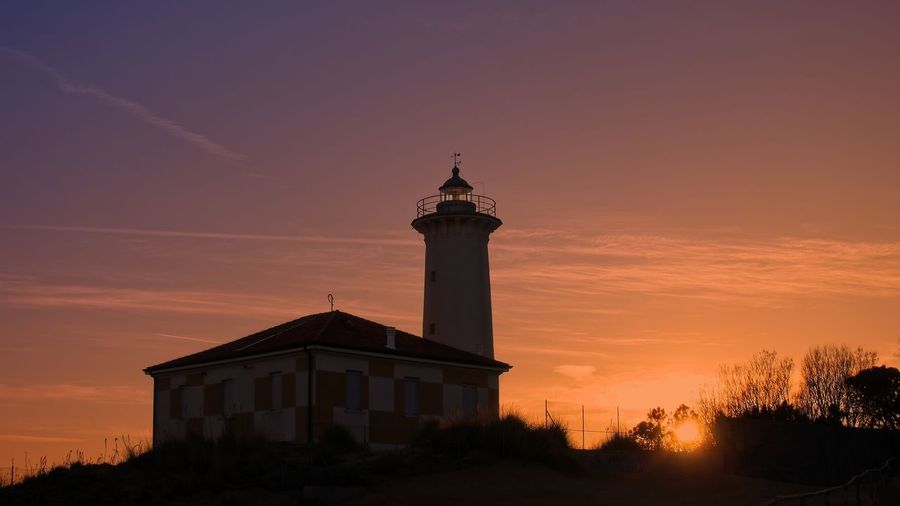 Silhouette lighthouse by building against sky during sunset