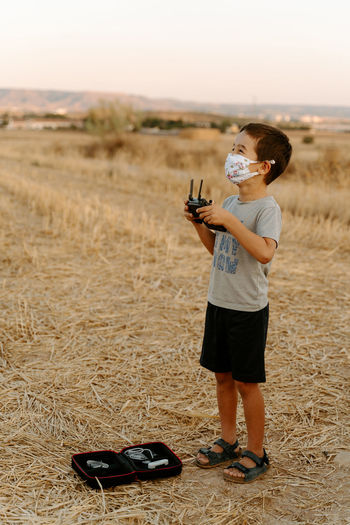 Little asian boy in protective mask using remote control while playing with flying drone in field