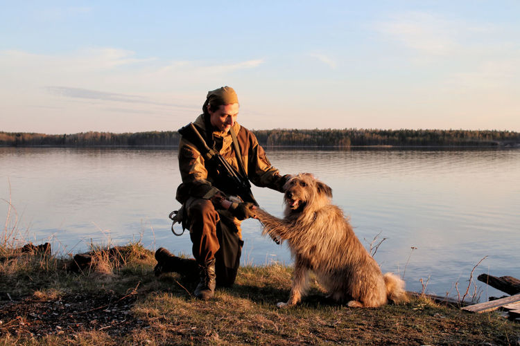 A man in a camouflage suit sitting on the shore of a lake with a dog