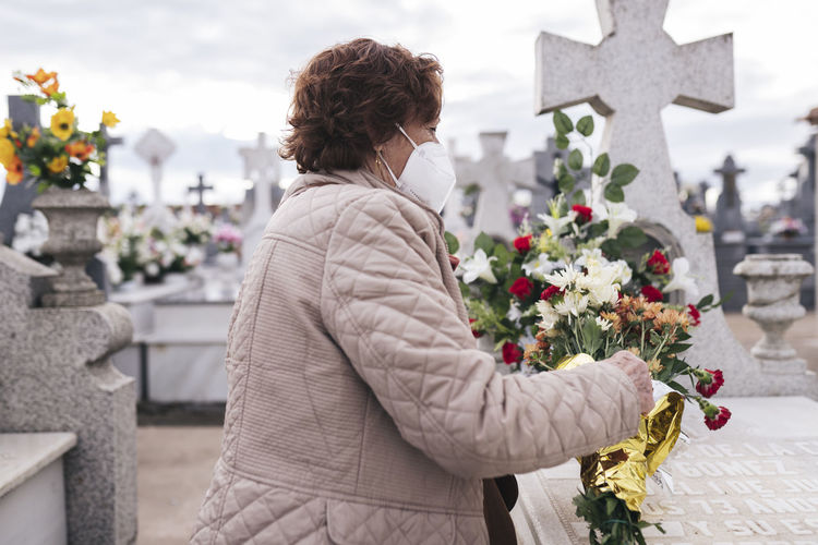 Senior woman wearing protective face mask putting flowers on tombstone in cemetery