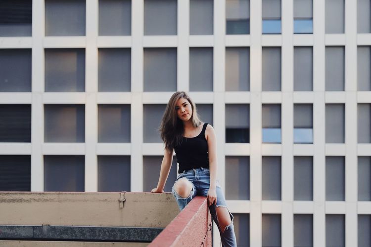 Portrait of young woman wearing torn jeans sitting on railing against modern building