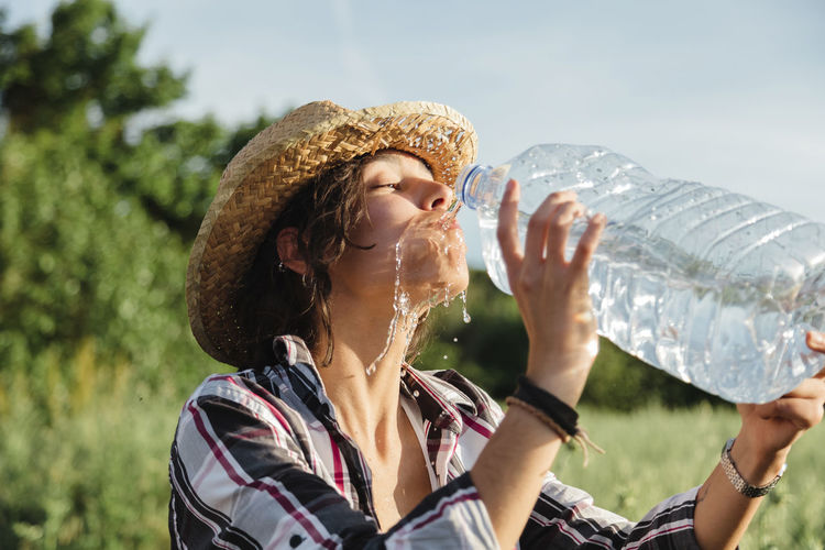 Young woman drinking water against trees and sky
