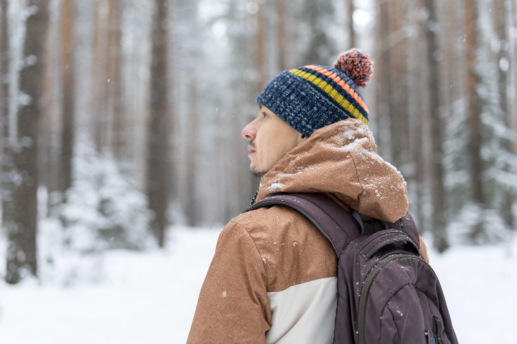 Rear view of young man in knitted hat with backpack walking in snowy forest in winter hiking 
