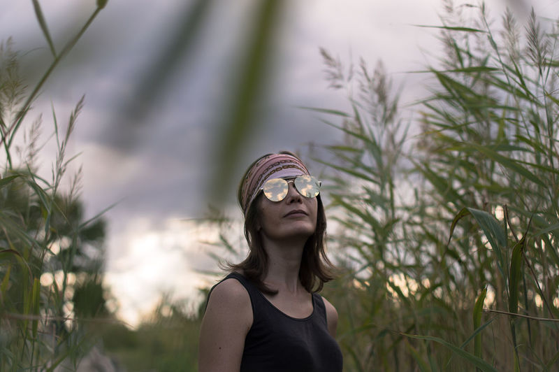 Mature woman wearing sunglasses standing amidst plants against cloudy sky during sunset