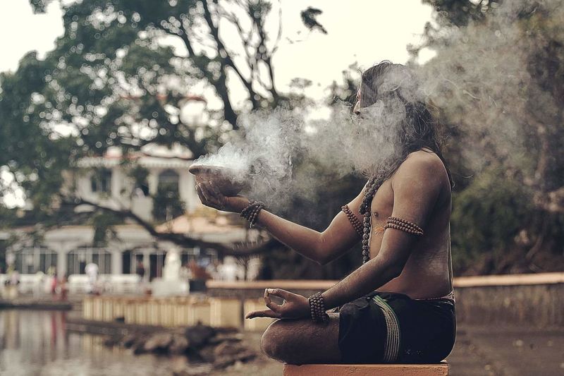 Side view of sadhu sitting with smoke emitting container on stool
