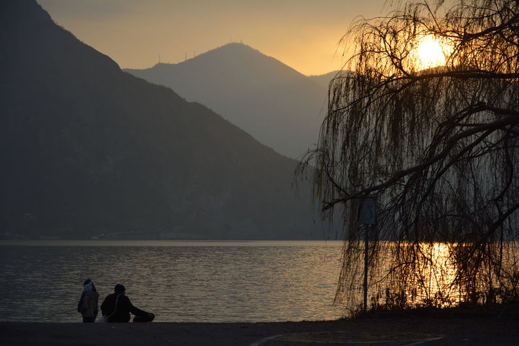 People on shore against silhouette mountains during sunset