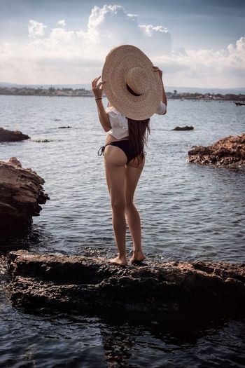 Rear view of woman wearing hat while standing on rock at beach against sky
