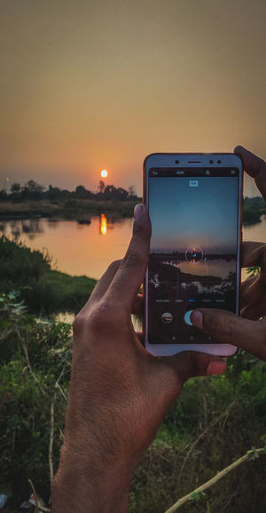 Cropped image of hand holding camera against sky during sunset