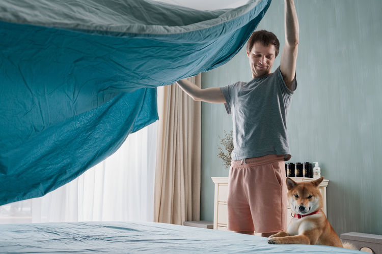 Young man and his funny dog are putting the bedding cover or mattress pad on the bed.