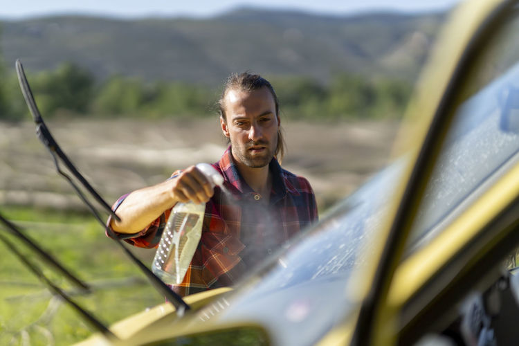 Man in checkered shirt spraying detergent on windscreen of van while washing vehicle during road trip in valley on sunny day