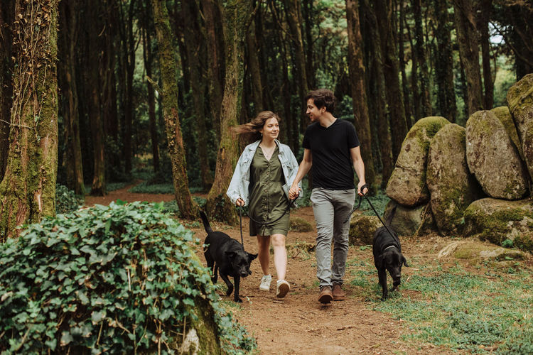 Couple with dogs on a leash smiling at each other, walking in forest
