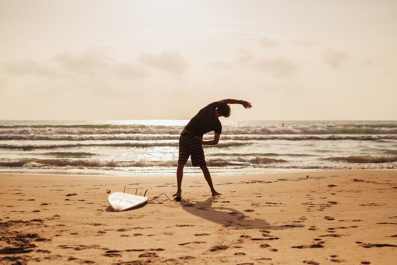 Surfer exercising on sand against sky during sunset