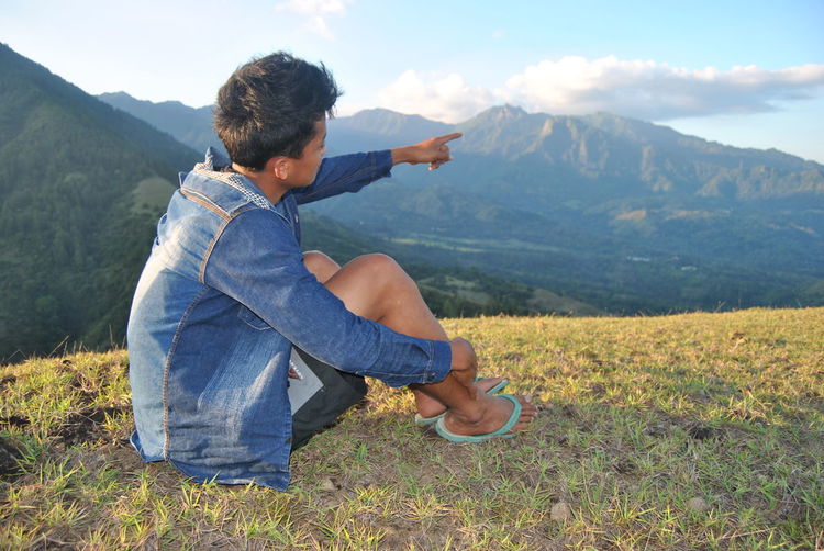 Young man pointing at mountain while sitting on cliff