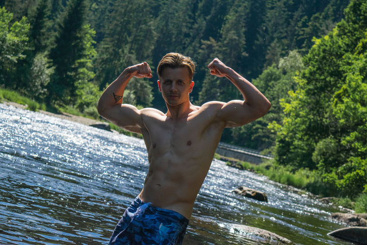 Portrait of shirtless man flexing muscles while standing by river in forest