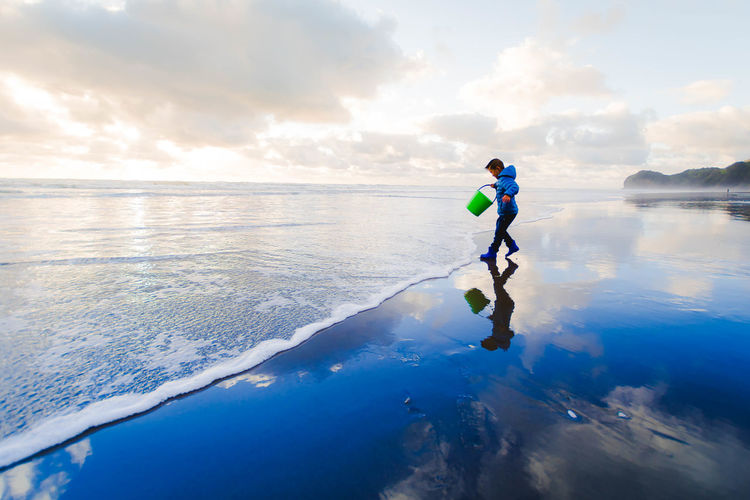 Side view of boy with bucket walking on shore at beach against sky