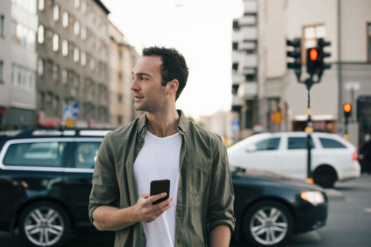 Man looking away while holding smart phone in city