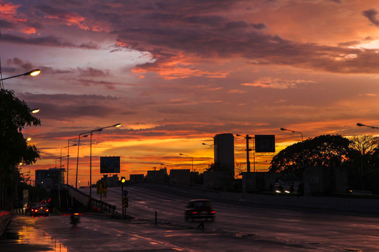 Road by illuminated city against orange sky