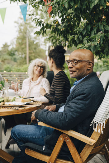 Smiling bald senior man sitting on chair enjoying dinner party by friends at back yard
