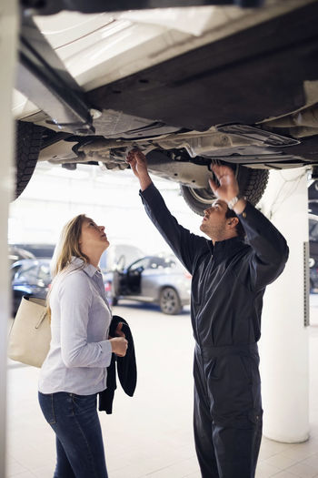 Mechanic with female customer standing under car at repair shop