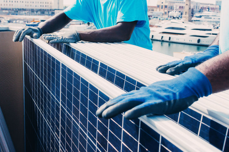 Crop unrecognizable african american male workers in protective gloves installing contemporary solar panels in industrial area of plant during daytime