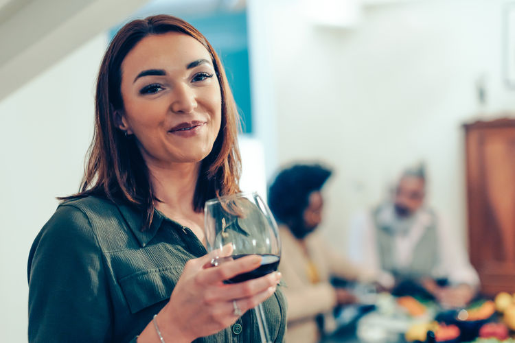 Happy lady looking in camera satisfied holding a glass of wine while friends talk in background