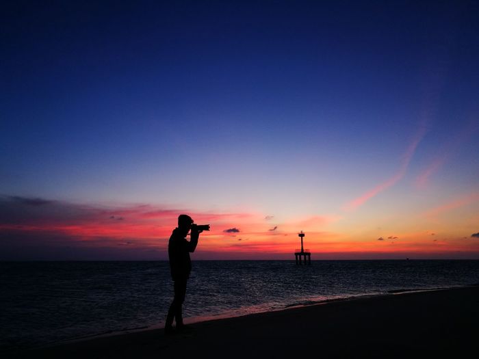 Silhouette woman photographing sea against sky during sunset