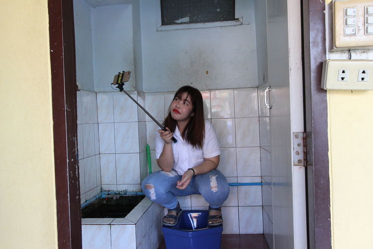 Young woman taking selfie on toilet bowl in bathroom at home