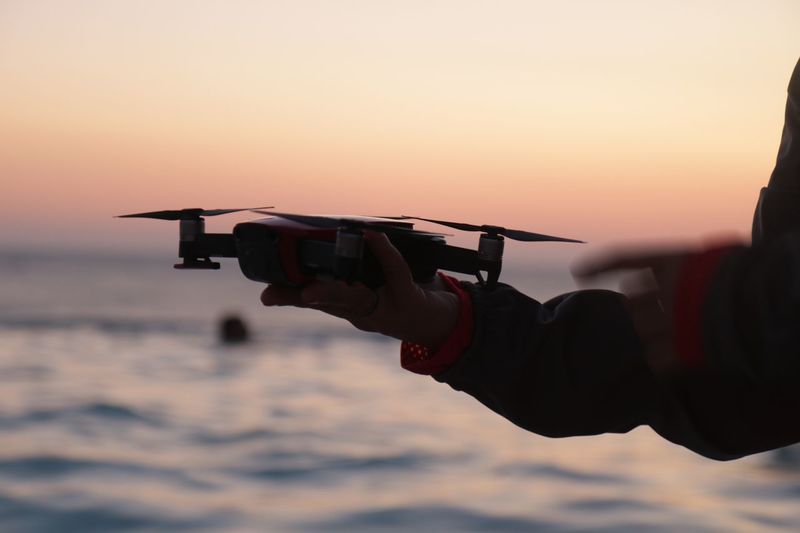 Cropped hand of woman holding drone against sea during sunset