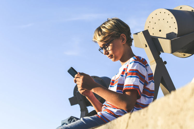 Low angle view of boy using phone while sitting against sky