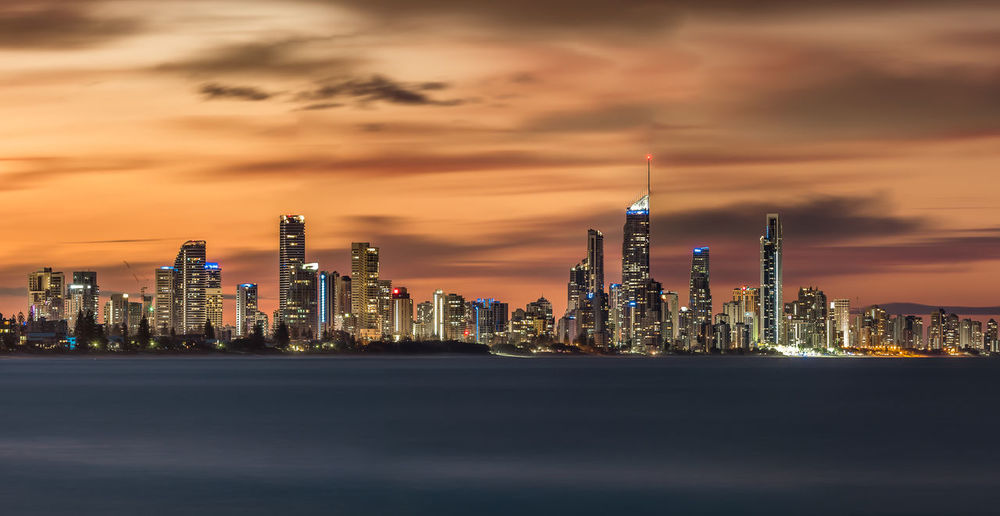 Illuminated modern buildings in city by sea against sky during sunset