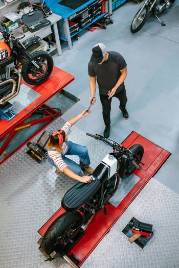 Unrecognizable mechanic giving tool to female coworker repairing motorbike on garaje