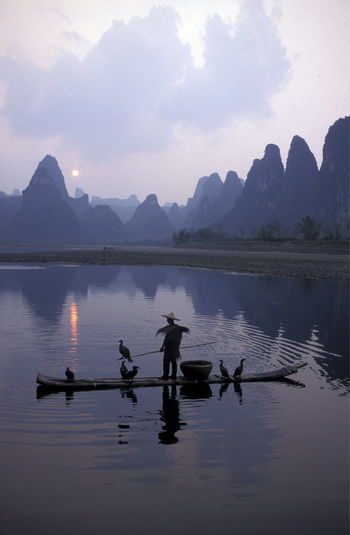 Man sailing boat with birds in river against mountains