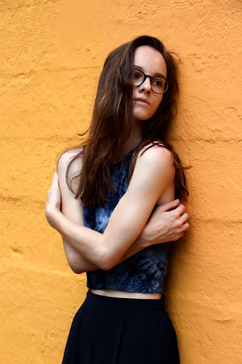 Thoughtful young woman with arms crossed standing by yellow wall