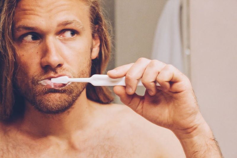 Close-up of shirtless man looking away while brushing teeth in bathroom