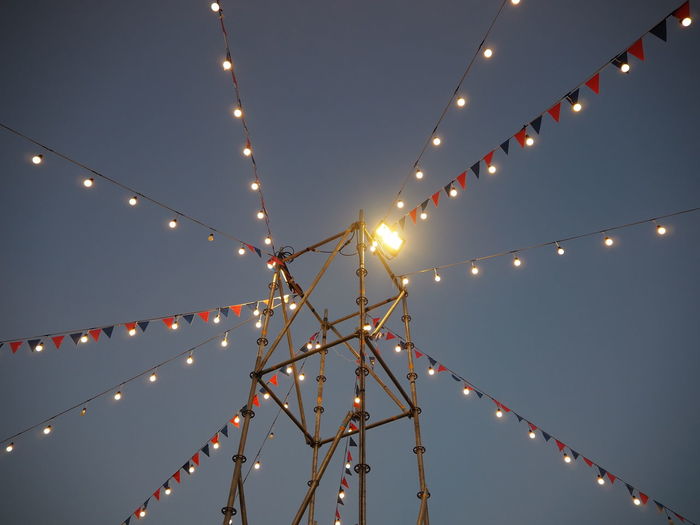 Low angle view of illuminated lighting equipment and buntings hanging against sky at dusk
