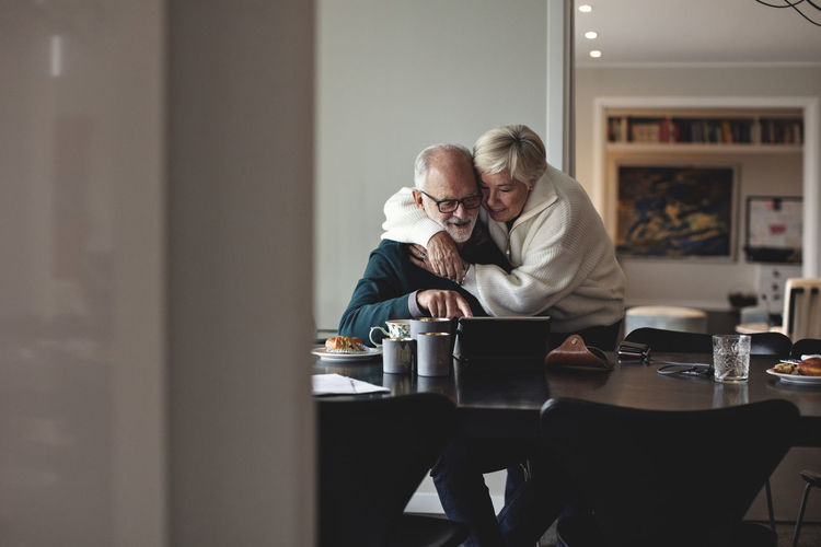 Smiling senior woman embracing partner by dining table in living room
