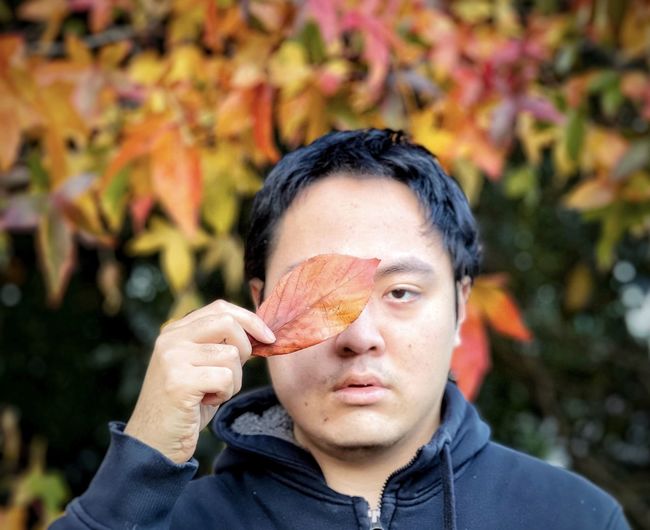 Portrait of man holding dried leaf against autumn foliage on trees.